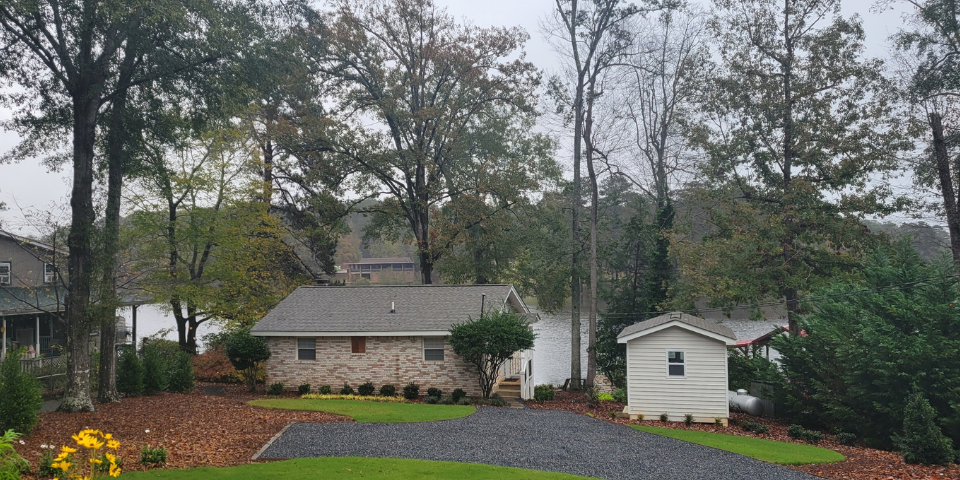 A lakeside property with a brick house, white shed, yellow flowers, tall trees, and a calm lake under an overcast sky.