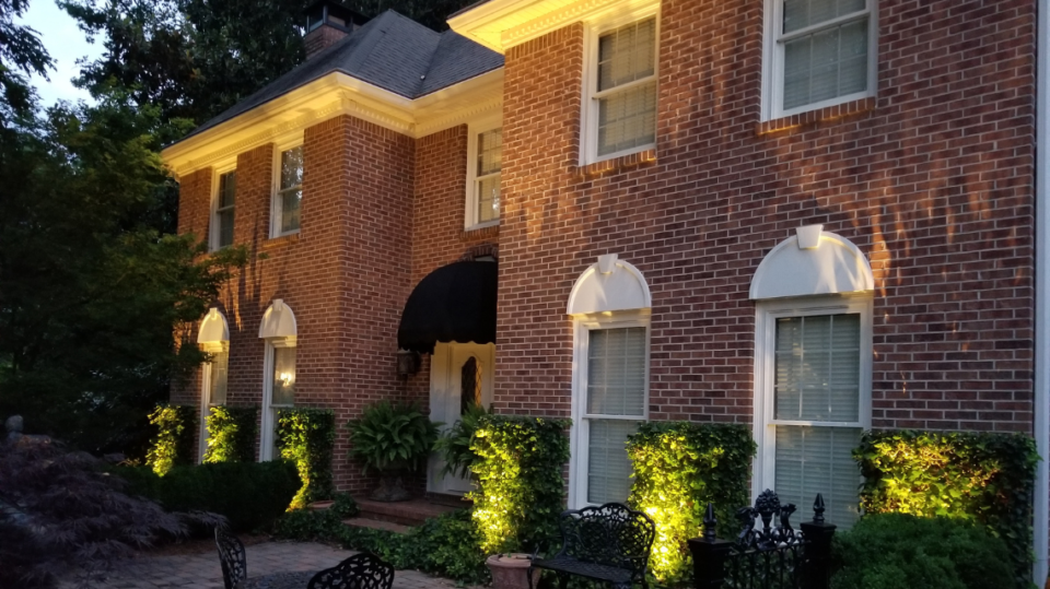 Elegant outdoor lighting highlights the brick façade of a two-story home in Fayette County, GA, with soft illumination on landscaping and architectural features, showcasing the expertise of a local landscaping company.